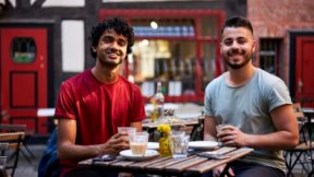 Zwei Studierende sitzen lächelnd draußen an einem Tisch im Café.